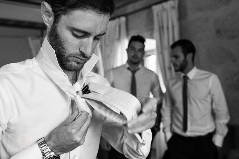 Novio haciendo nudo de la corbata en habitacion con los amigos - Fotografía de boda